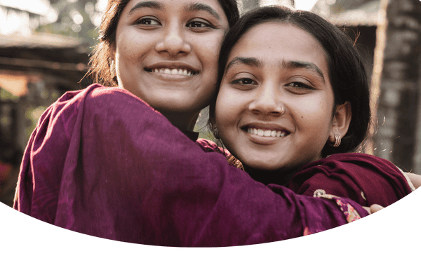 Two girls hugging and smiling at the camera in a sunny outdoor setting.