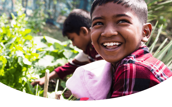 Smiling boy in a red and black plaid shirt crouching in a garden, with another boy in the background picking vegetables.